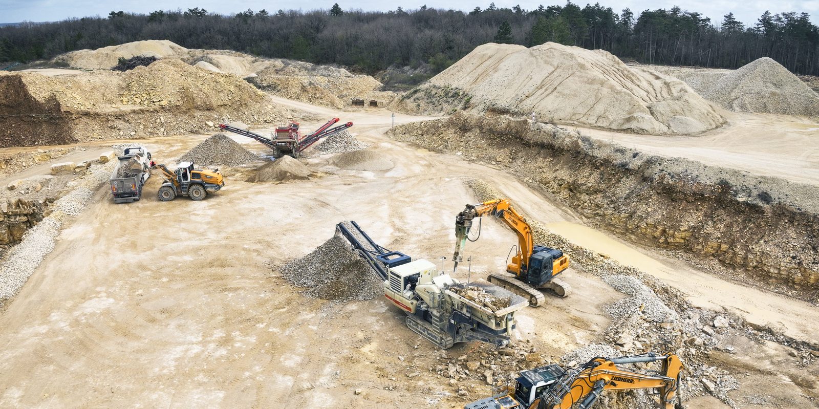 Chantier vu par drone : pelles hydrauliques, concasseur mobile, crible, chargeuse et camion au milieu des stocks de matériaux